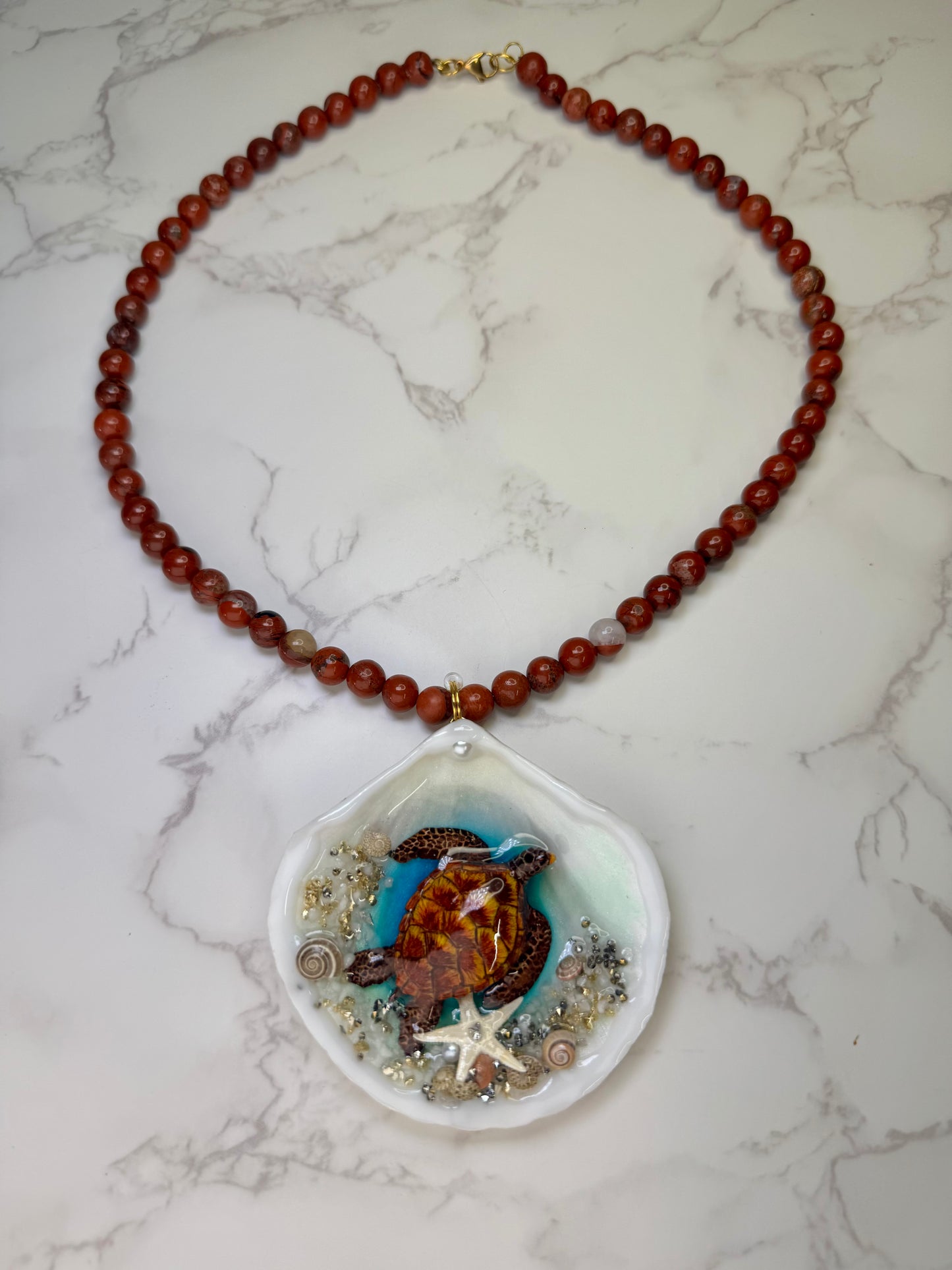Golden Ember Current Natural seashell featuring a beautiful metal handmade metal brown-and-yellow sea turtle, sand, golden sparkles, seashells, and a tiny starfish, preserved in clear UV resin paired with a crimson crystal ball necklace