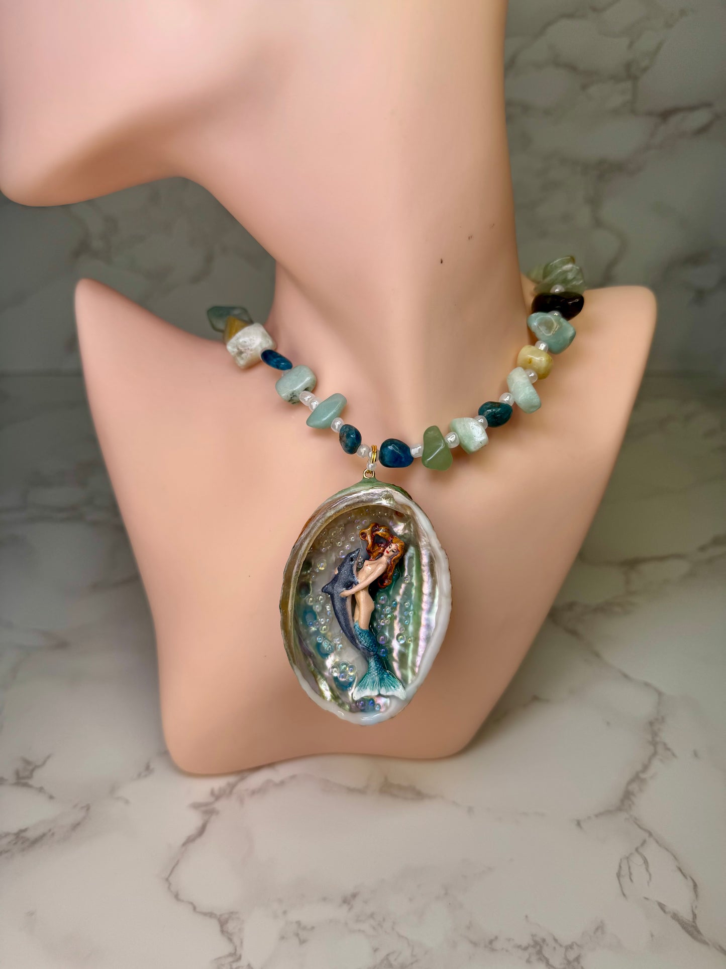 Azure Harmony Siren Natural seashell featuring a miniature mermaid with orange hair and a shimmering blue tail holding a blue dolphin