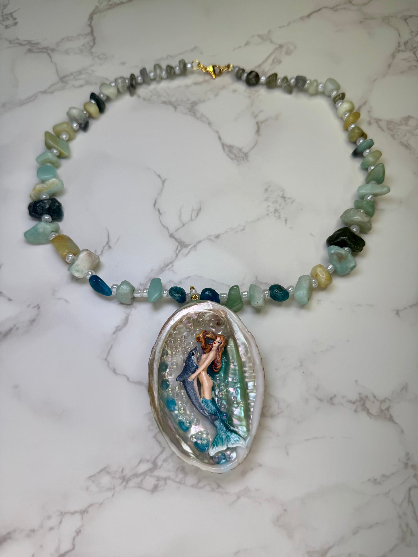 Azure Harmony Siren Natural seashell featuring a miniature mermaid with orange hair and a shimmering blue tail holding a blue dolphin