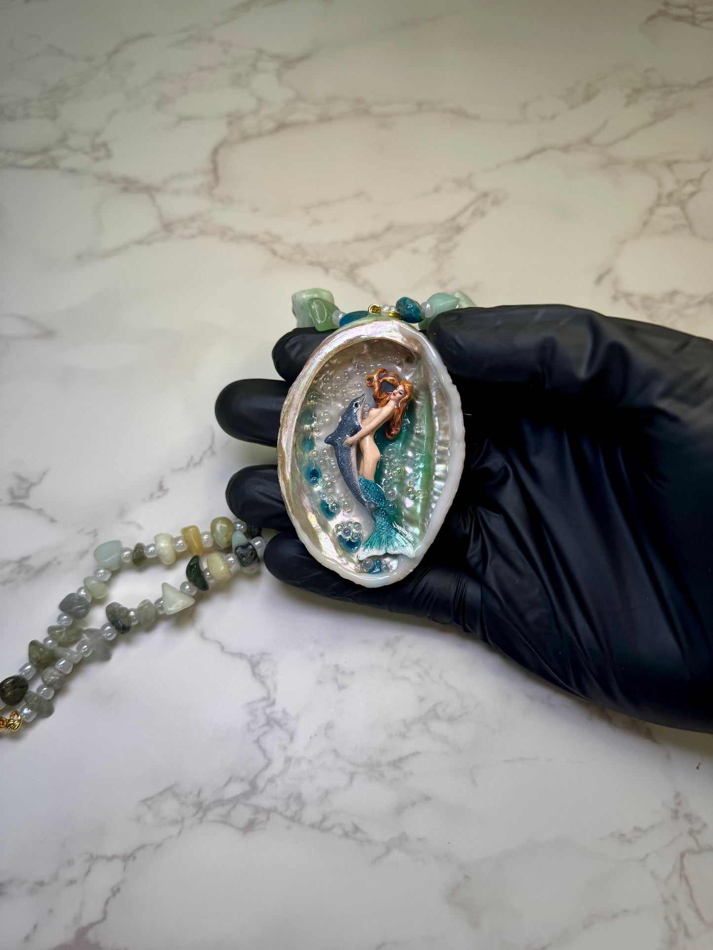 Azure Harmony Siren Natural seashell featuring a miniature mermaid with orange hair and a shimmering blue tail holding a blue dolphin